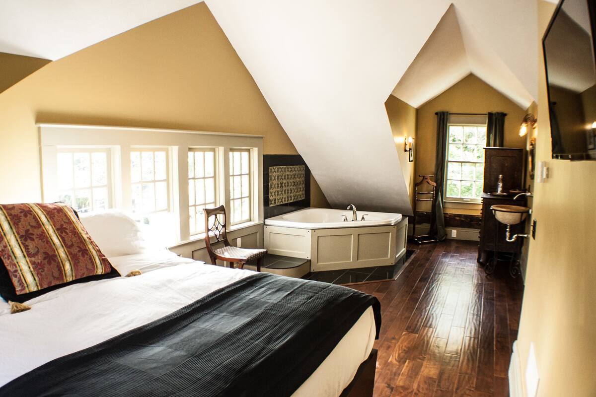 A bedroom with a large bed, a sitting area with a chair, a bathtub, and a small sink.