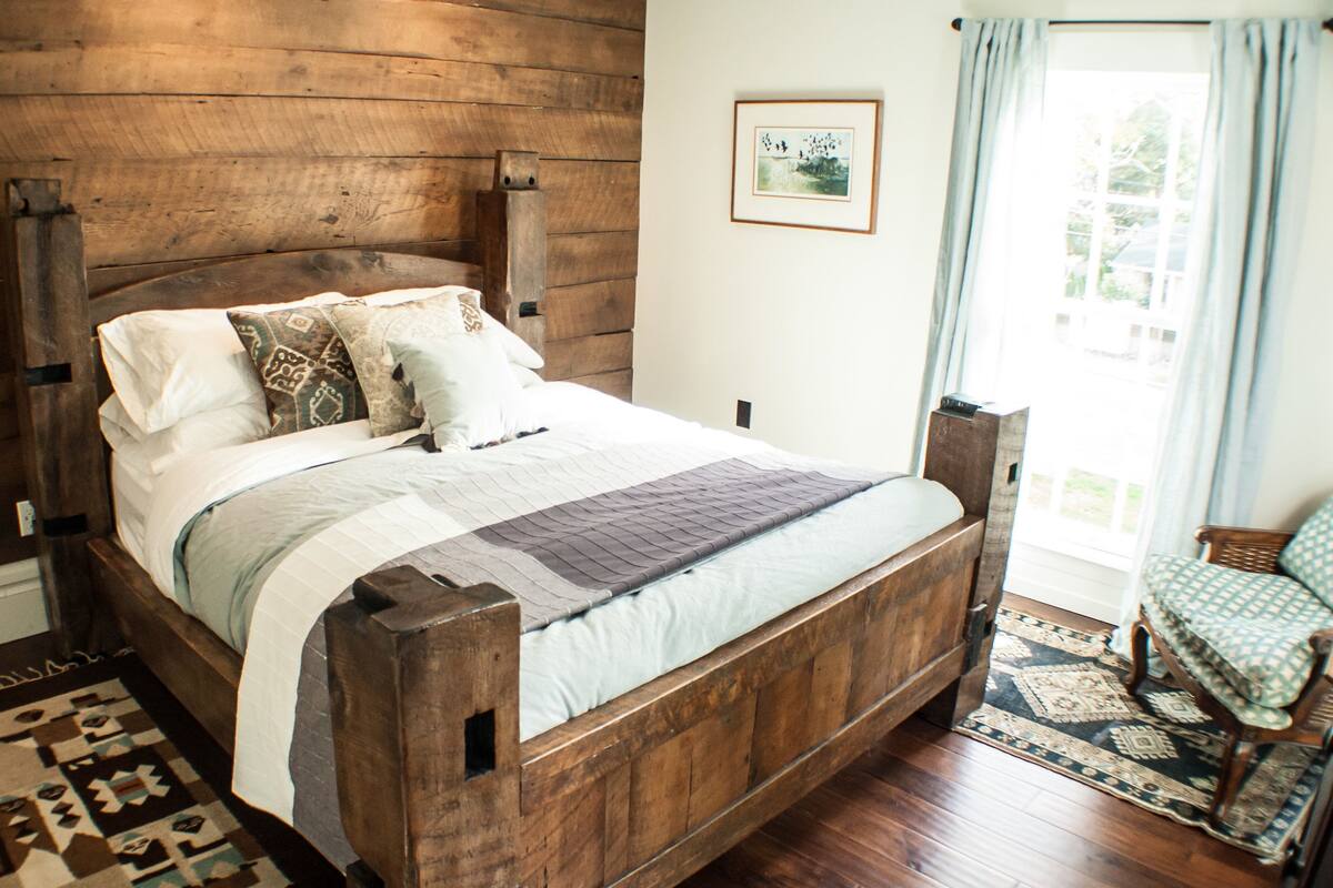 A wooden bed with a quilt, pillows, and a rug in a room with a wooden wall and a window with curtains.