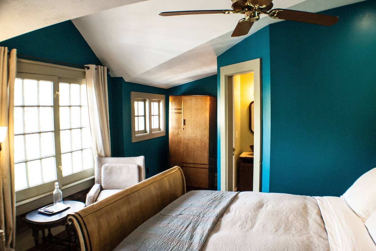 A hotel room with a bed, a desk, a chair, a wardrobe, and a ceiling fan.