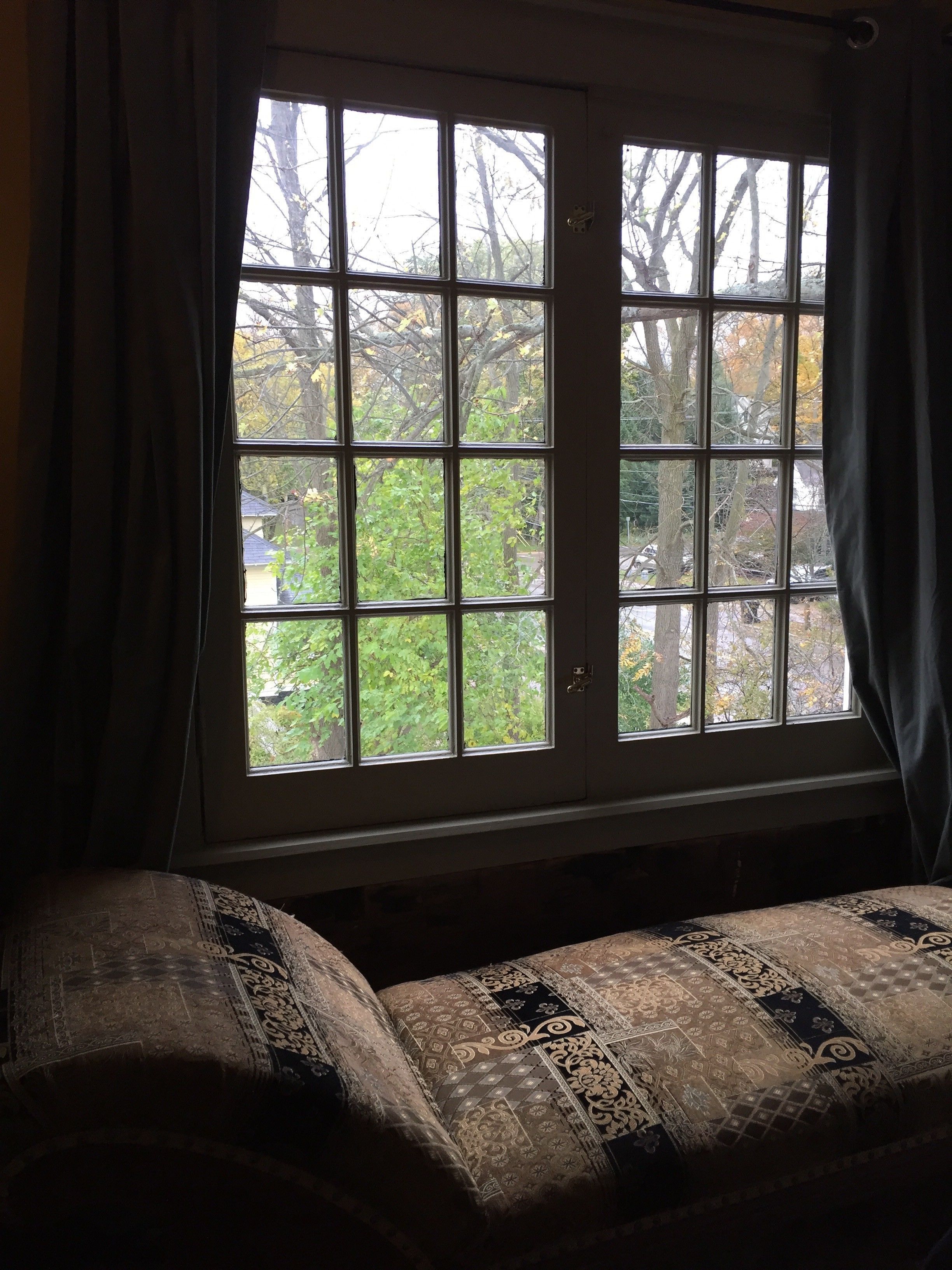 Una habitación con un ventanal grande, una cama con ropa de cama estampada y vistas a árboles y una casa.