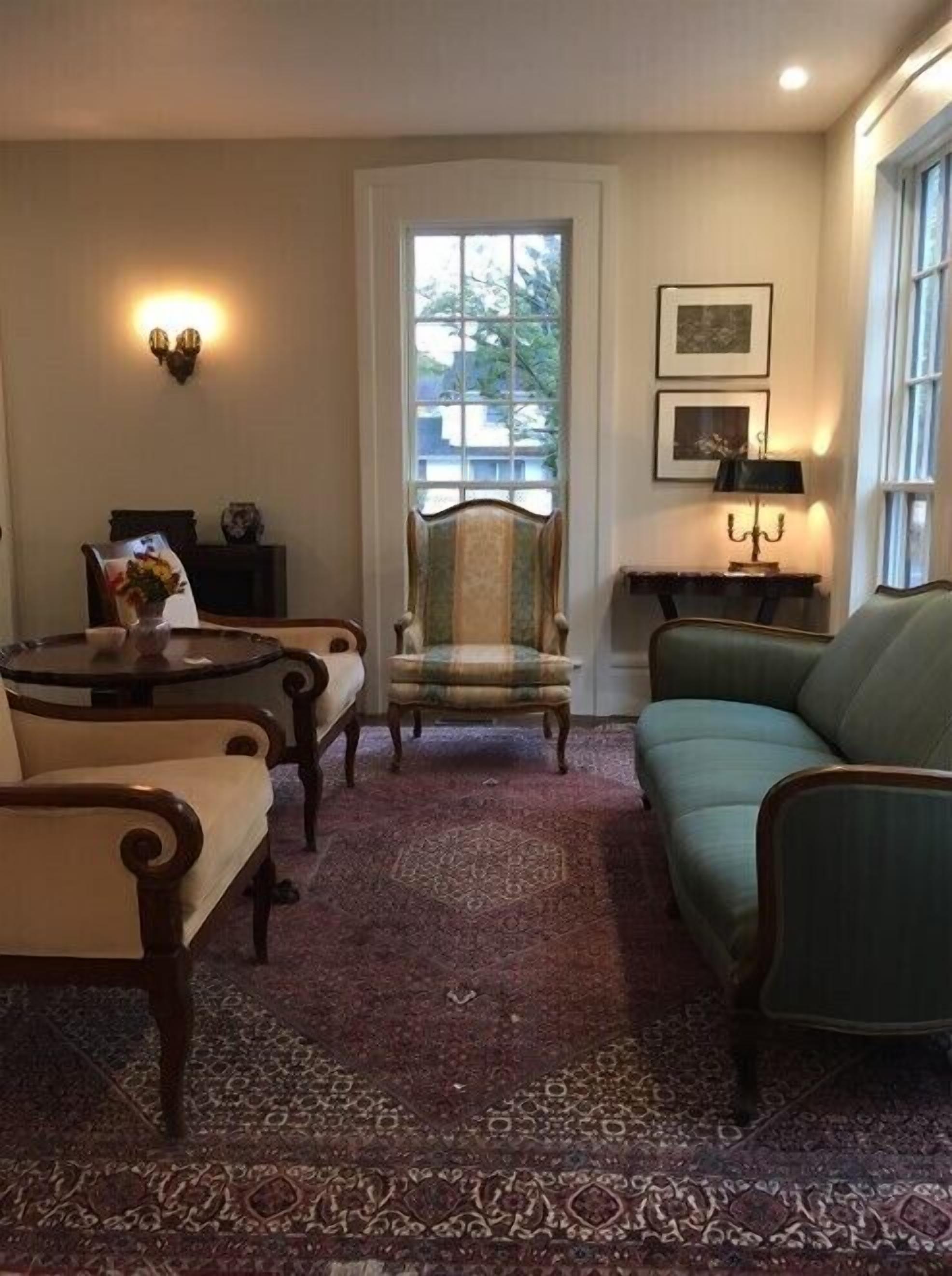 Een woonkamer met een groene bank, twee fauteuils, een houten salontafel, een tapijt met een patroon, een stoel met een ruitkussen, en een deur met een raam.