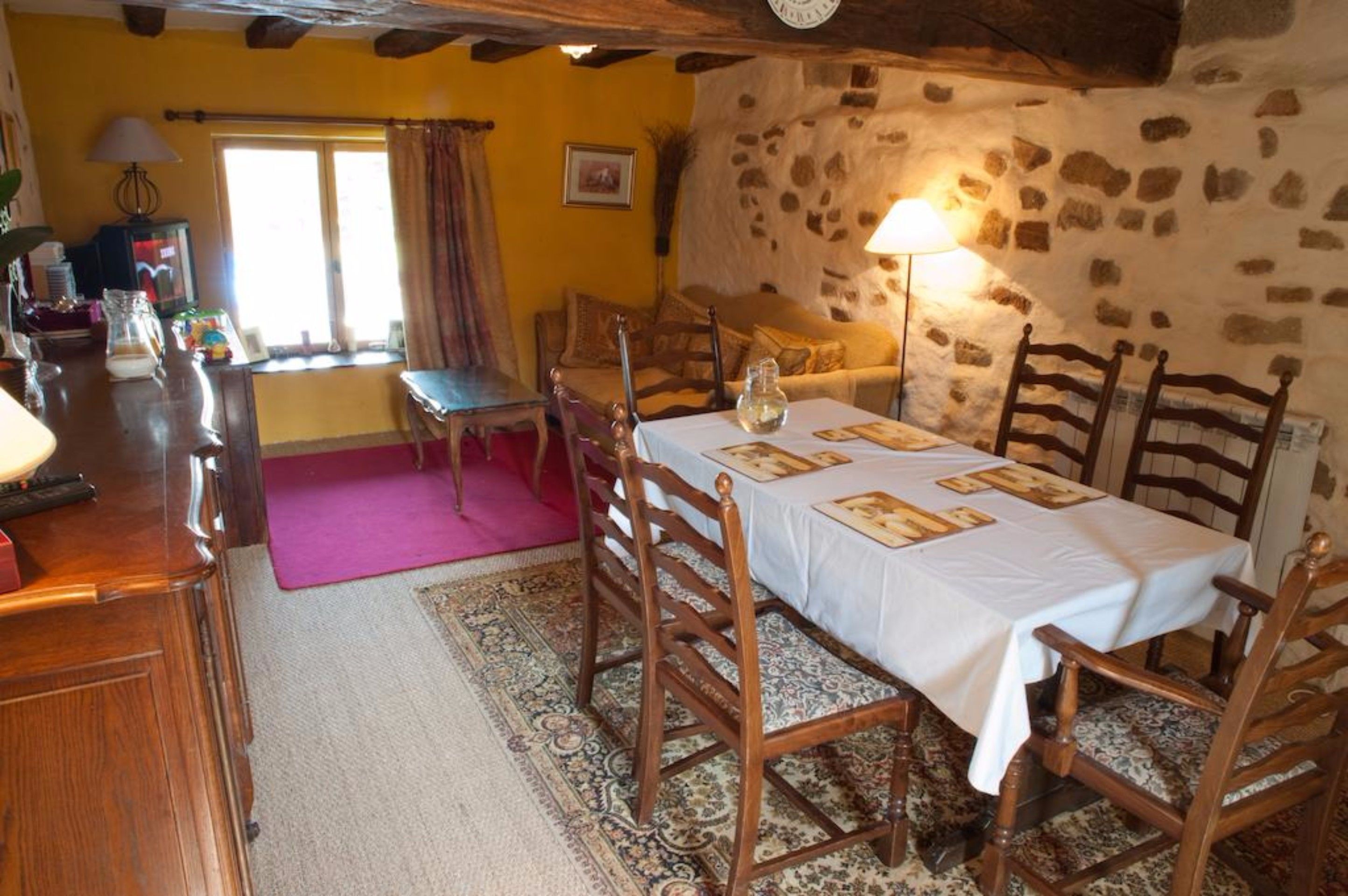 A dining area with a table set for four, surrounded by wooden chairs. A stone wall and a window with curtains are visible in the background.