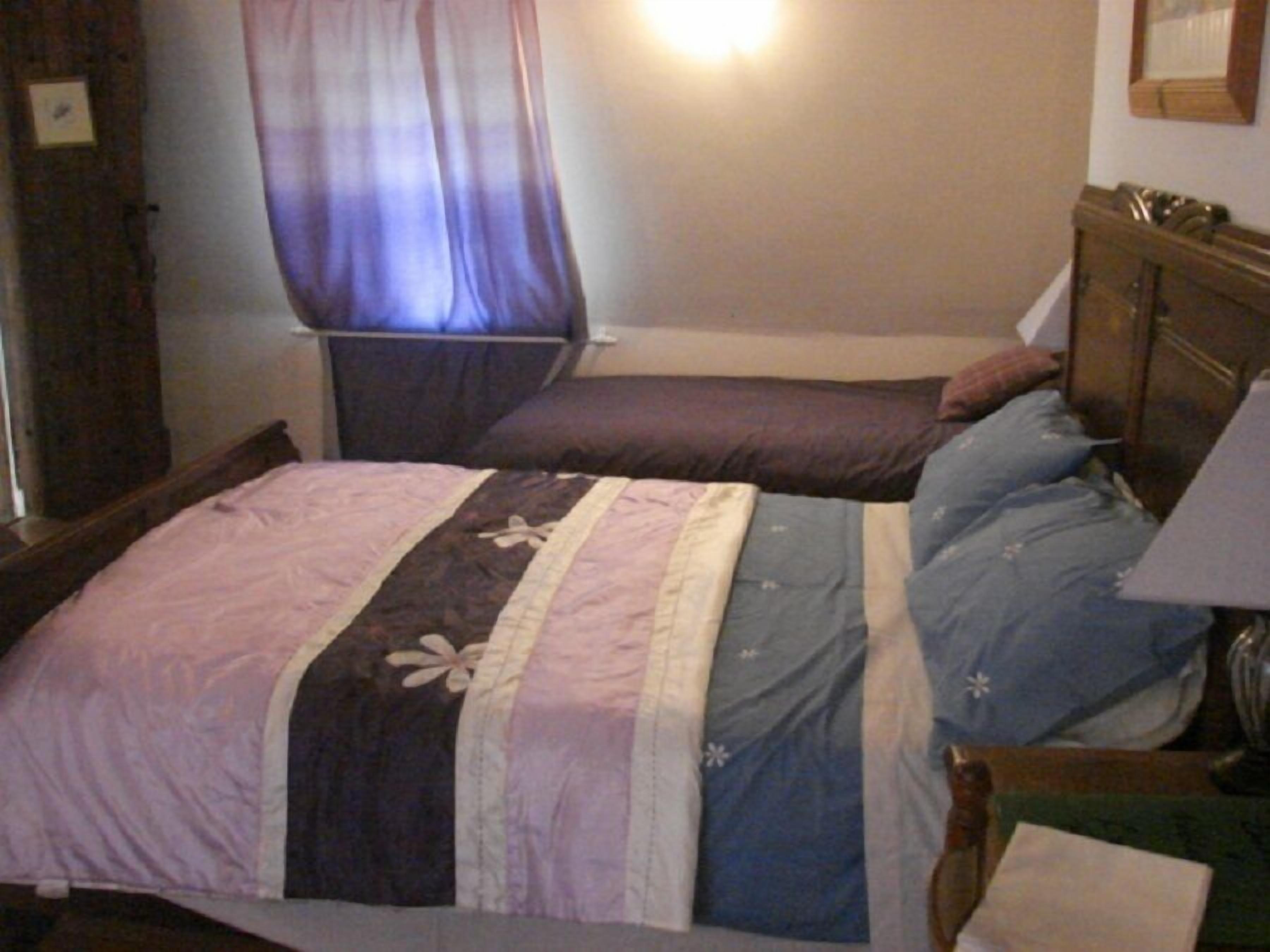 A bedroom with two beds, a wooden headboard, a bedside table with a lamp, and a window with curtains.