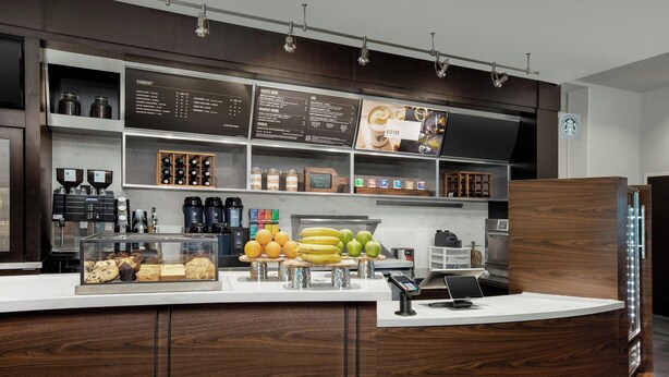 A modern coffee shop interior with a counter displaying pastries and fruits, a menu board, and coffee machines.