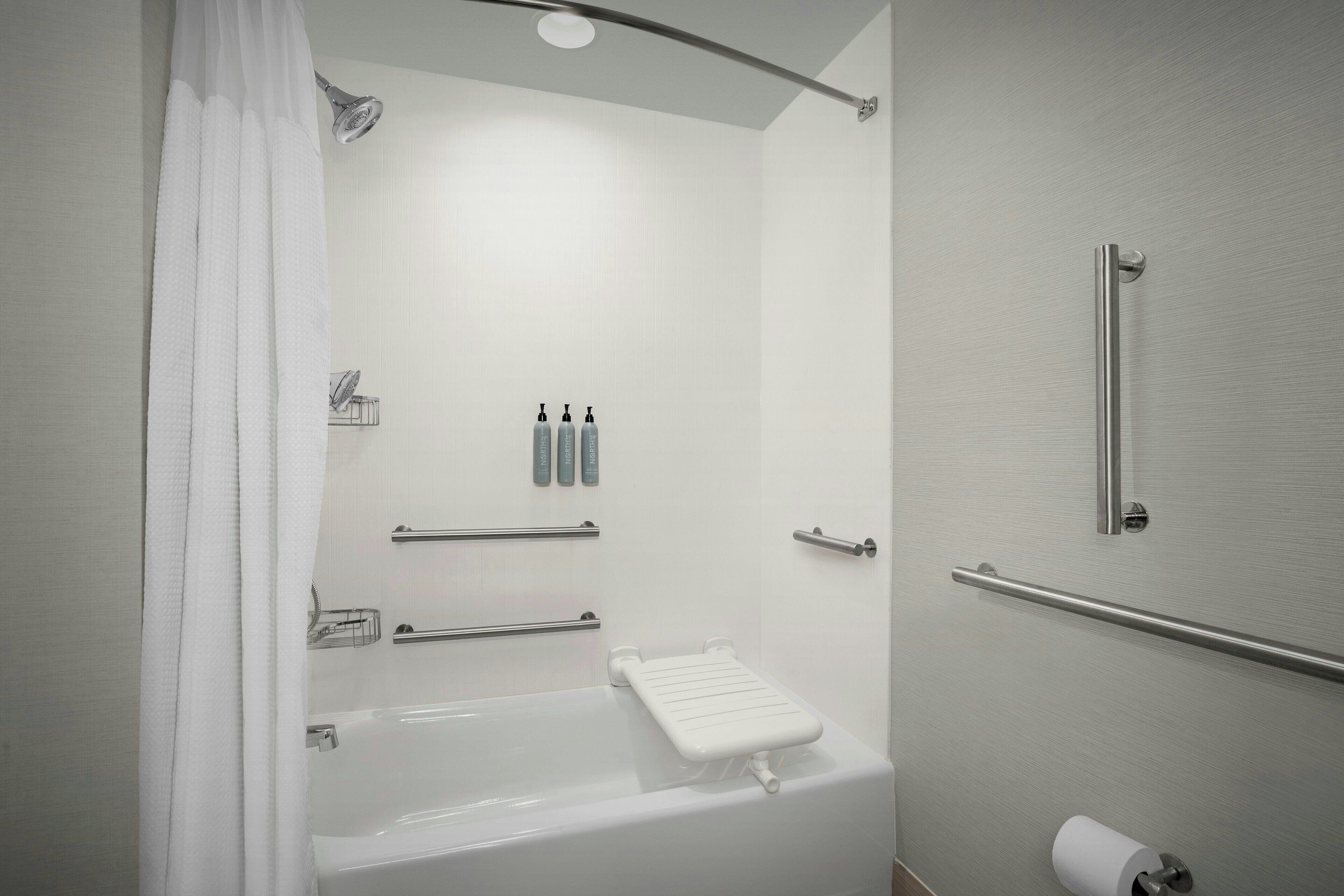 Un baño moderno con bañera, ducha e inodoro. Hay barras de apoyo, un perchero para toallas y un dispensador de papel higiénico.
