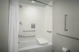 Un baño moderno con bañera, ducha e inodoro. Hay barras de apoyo, un perchero para toallas y un dispensador de papel higiénico.