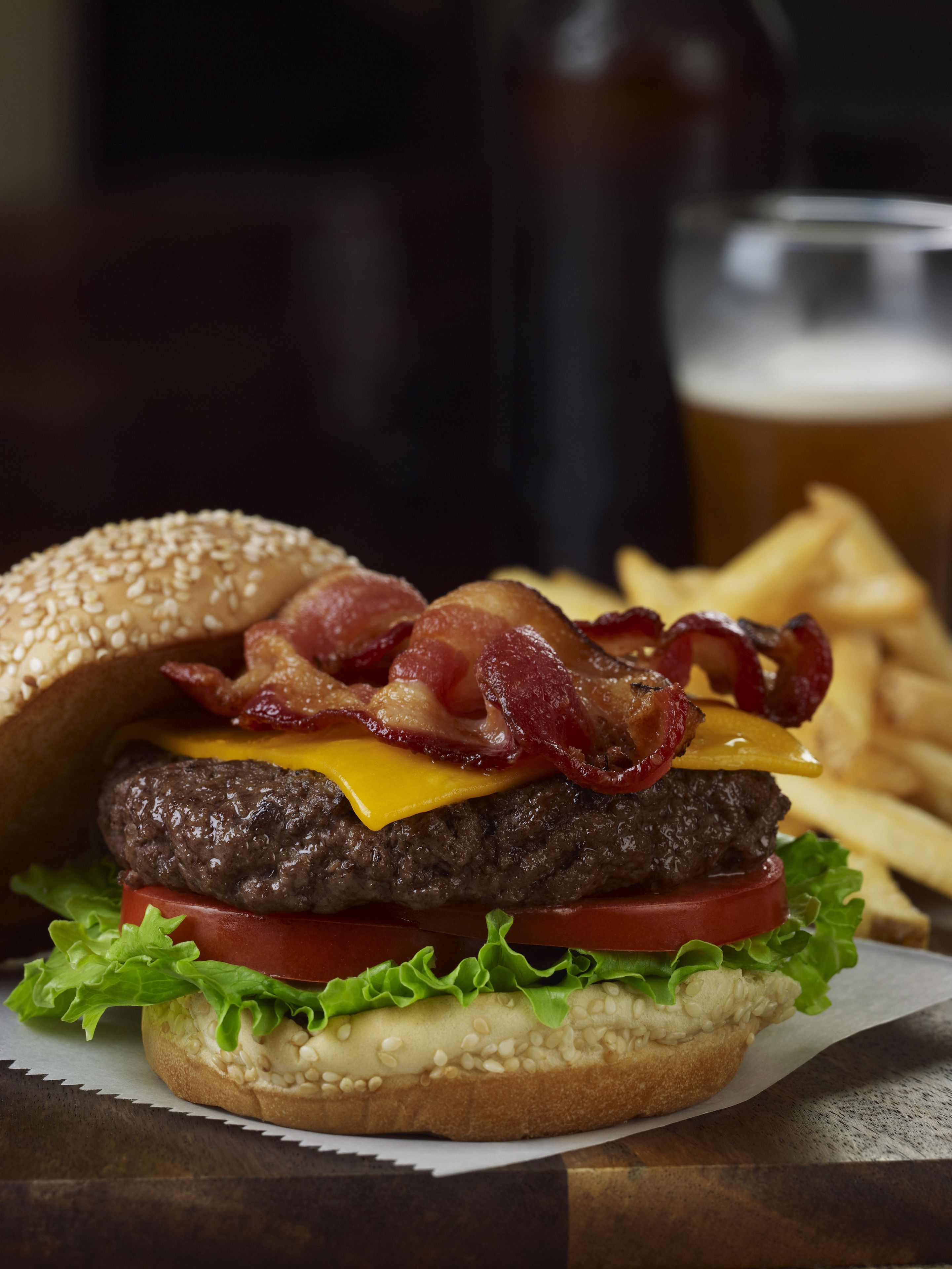 A burger with bacon, cheese, and lettuce on a bun, accompanied by fries and a glass of beer.