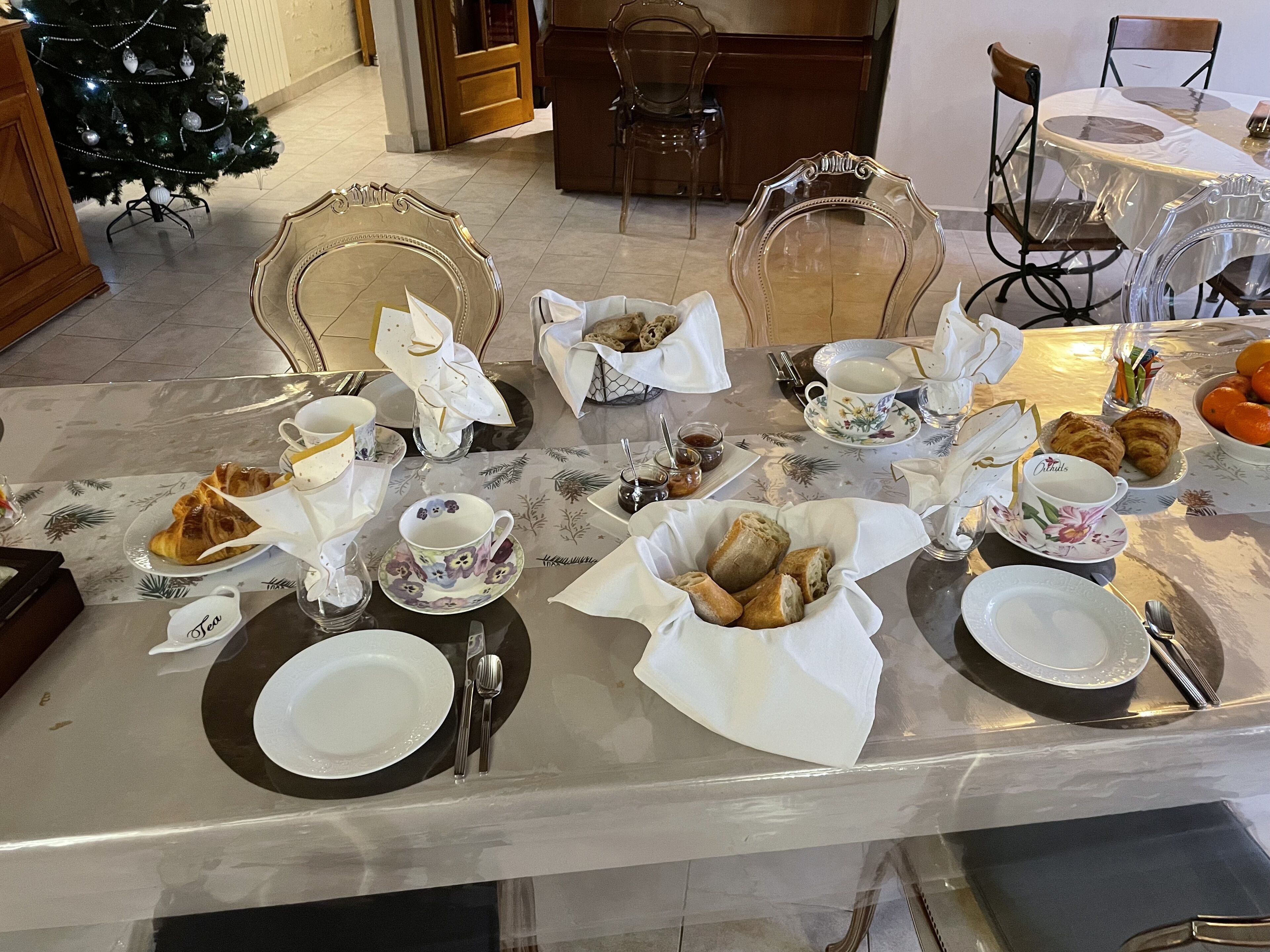 A dining table set with plates, cups, and pastries, surrounded by chairs and a Christmas tree.