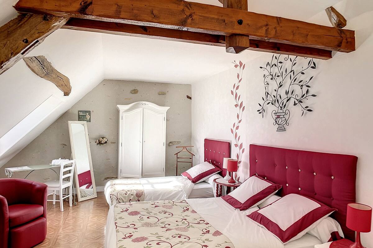 A bedroom with a sloped ceiling, a bed with red and white bedding, a red armchair, a small table with a red lamp, and a patterned rug.