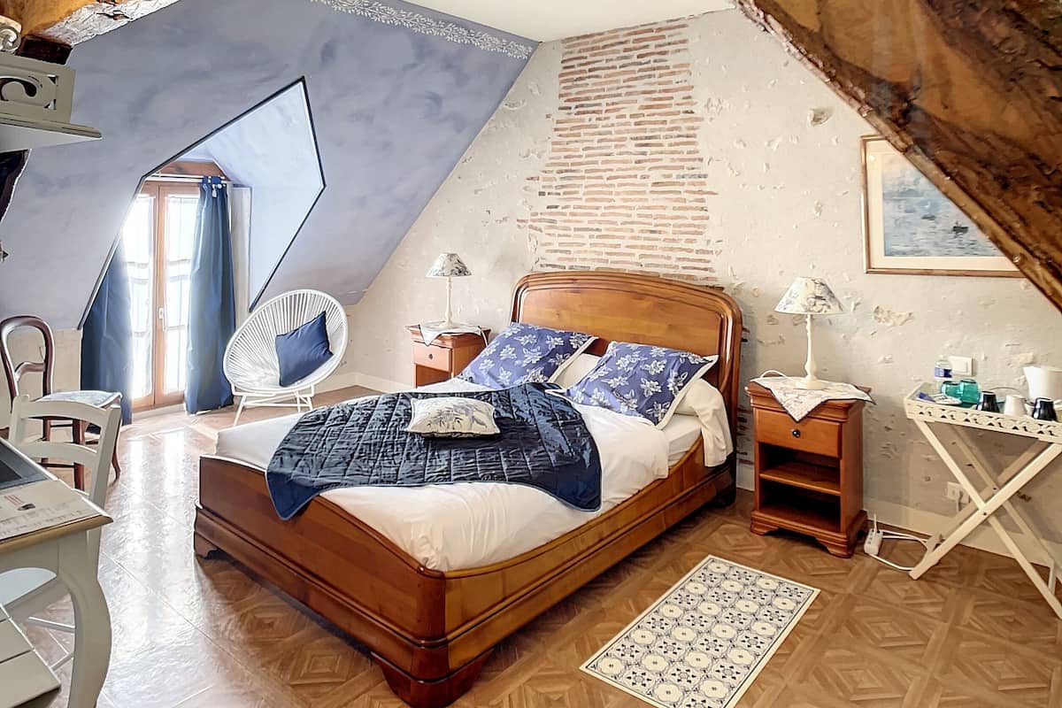 A bedroom with a sloped ceiling, a wooden bed, a chair, a desk, and a small table with a lamp.