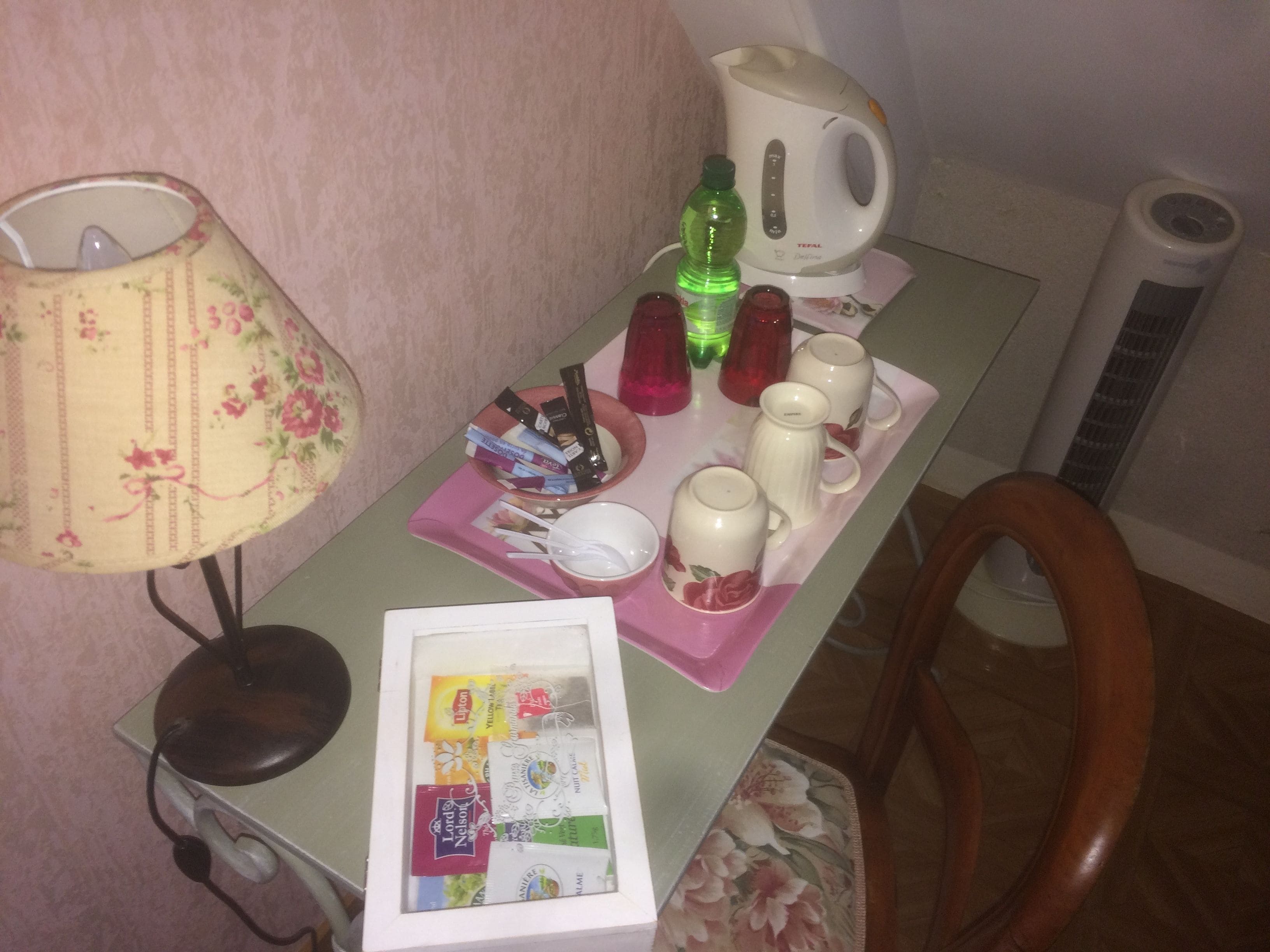 Een kleine tafel met een lamp, een waterkoker, een dienblad met kopjes en een schaal, en een doosje theezakjes.