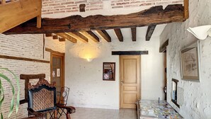 Ein geräumiger Raum mit hoher Decke, freiliegenden Holzbalken, einer Ziegelwand und einer Holztreppe. Ein Holztisch ist mit Zeitschriften, einem Sessel und einer Topfpflanze bestückt.
