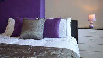 A neatly made bed with a white and brown comforter, a purple and white pillow arrangement, and a bedside table with a lamp.