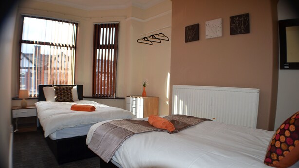 A hotel room with two beds, a nightstand, a lamp, a radiator, and a window with blinds. - Birchfields Guest House (Manchester)