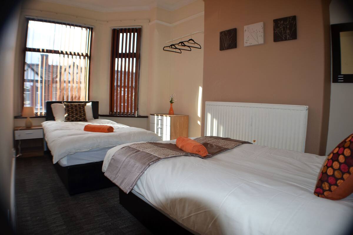 A hotel room with two beds, a nightstand, a lamp, a radiator, and a window with blinds.