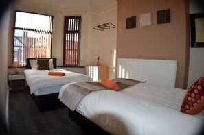A hotel room with two beds, a nightstand, a lamp, a radiator, and a window with blinds. - Birchfields Guest House (Manchester)
