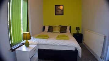 A neatly made bed with pillows and a green towel, a bedside table with a lamp, and a radiator on the wall.