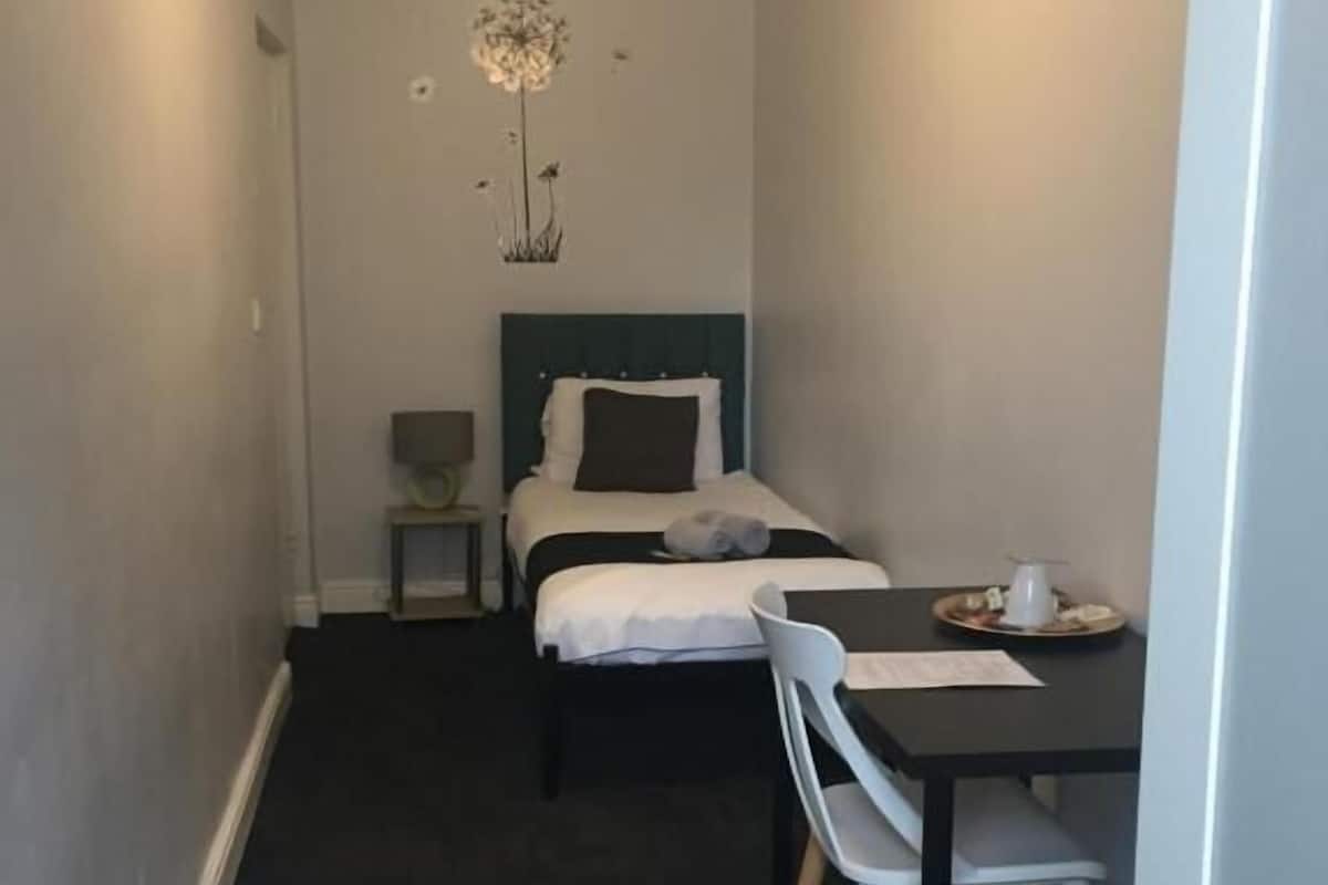 A narrow hotel room with a single bed, a small table with a chair, and a hanging light fixture.