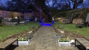 Un vialetto in pietra conduce a una pergola in legno con luci a stringa, circondata da fioriere con fiori gialli.