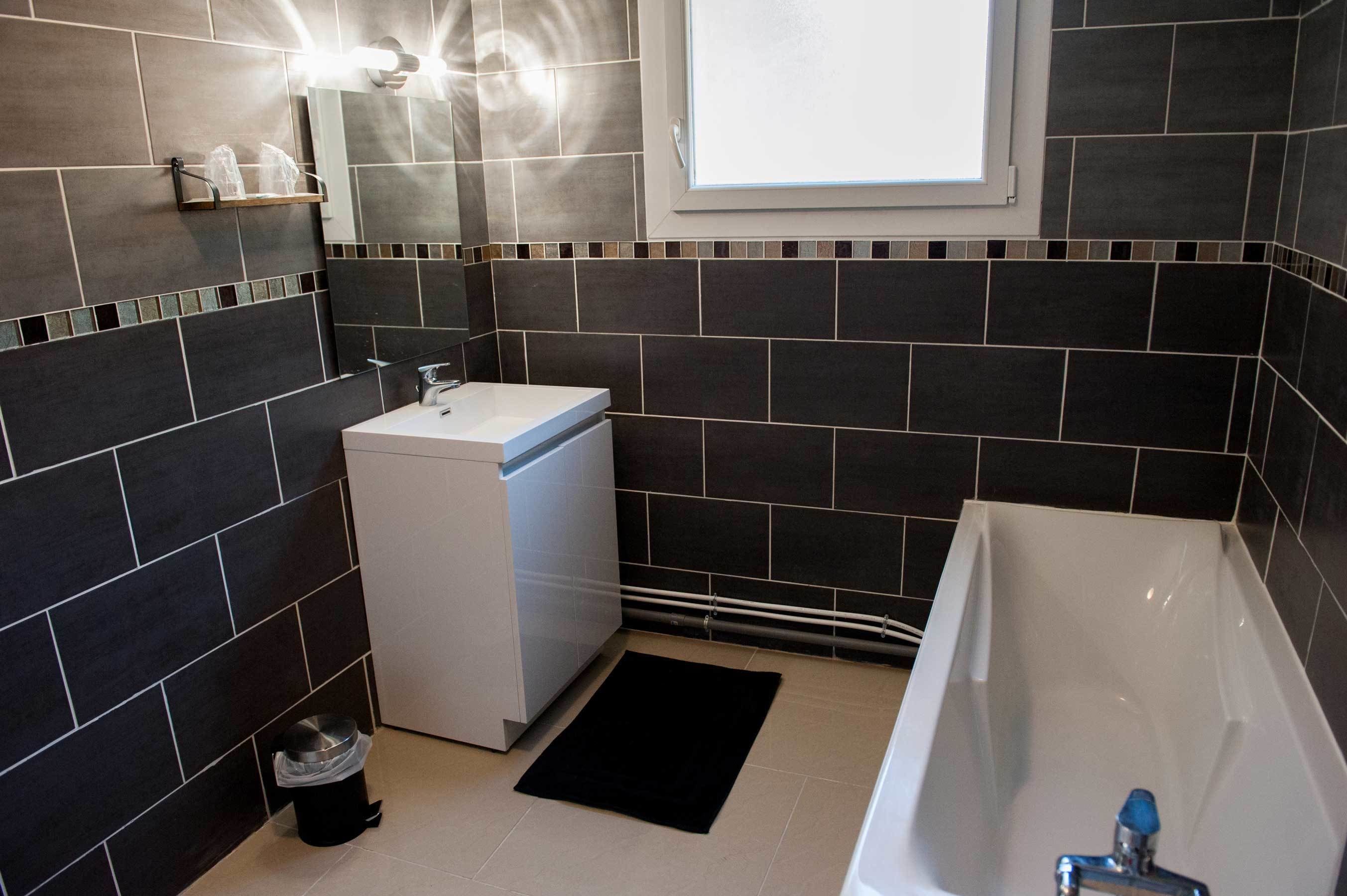 A modern bathroom with dark tiles, a white sink, a compact washing machine, and a bathtub.