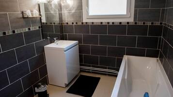 A modern bathroom with dark tiles, a white sink, a compact washing machine, and a bathtub.