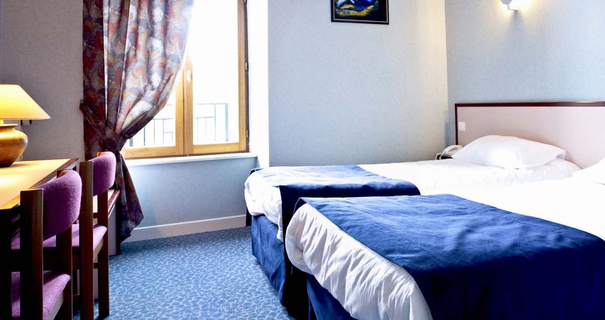 A hotel room with two beds, a desk with a lamp, and a window with curtains.