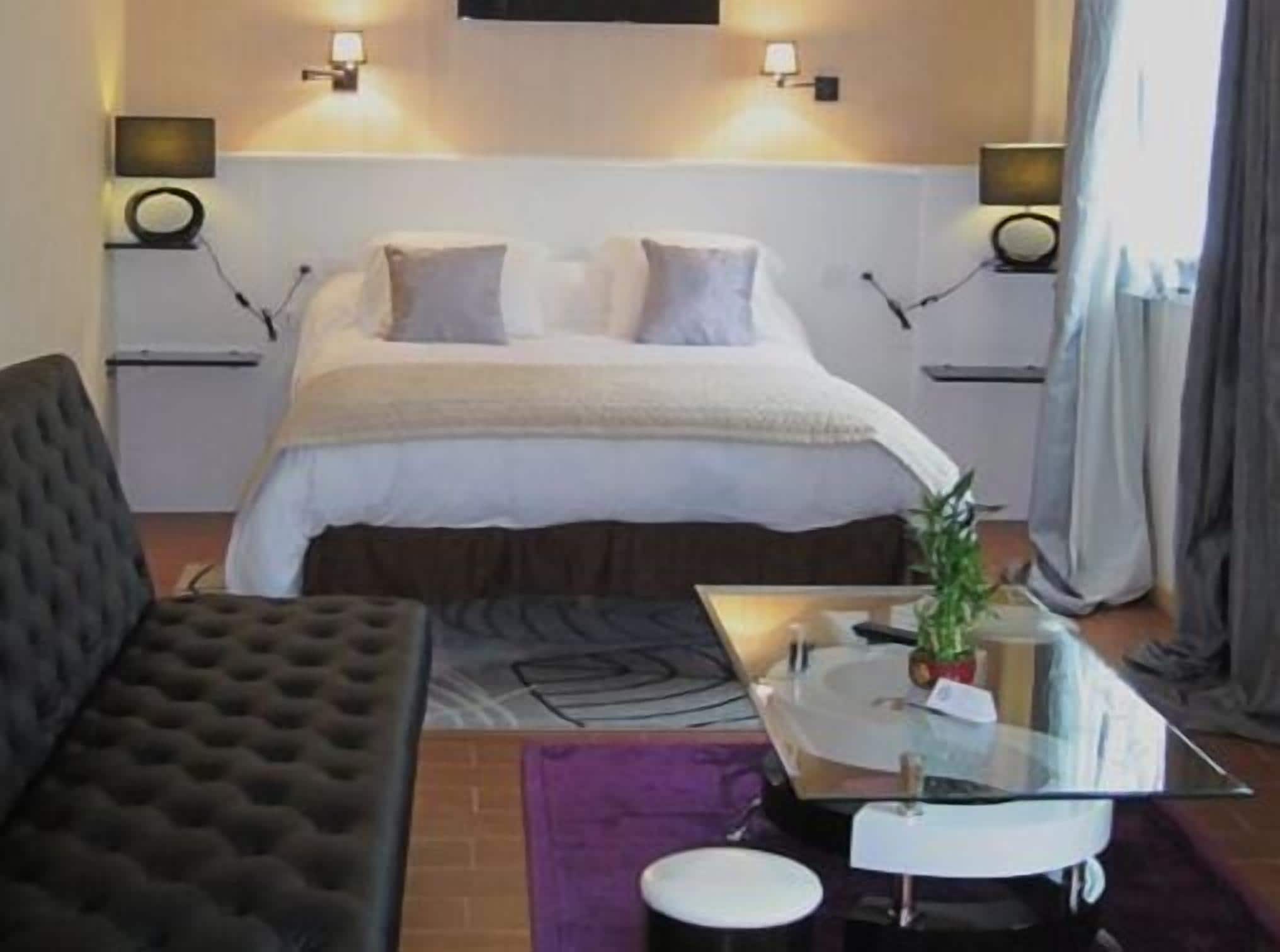 Quarto de hotel com cama, sofá, mesa de centro de vidro e uma pequena mesa com uma planta.