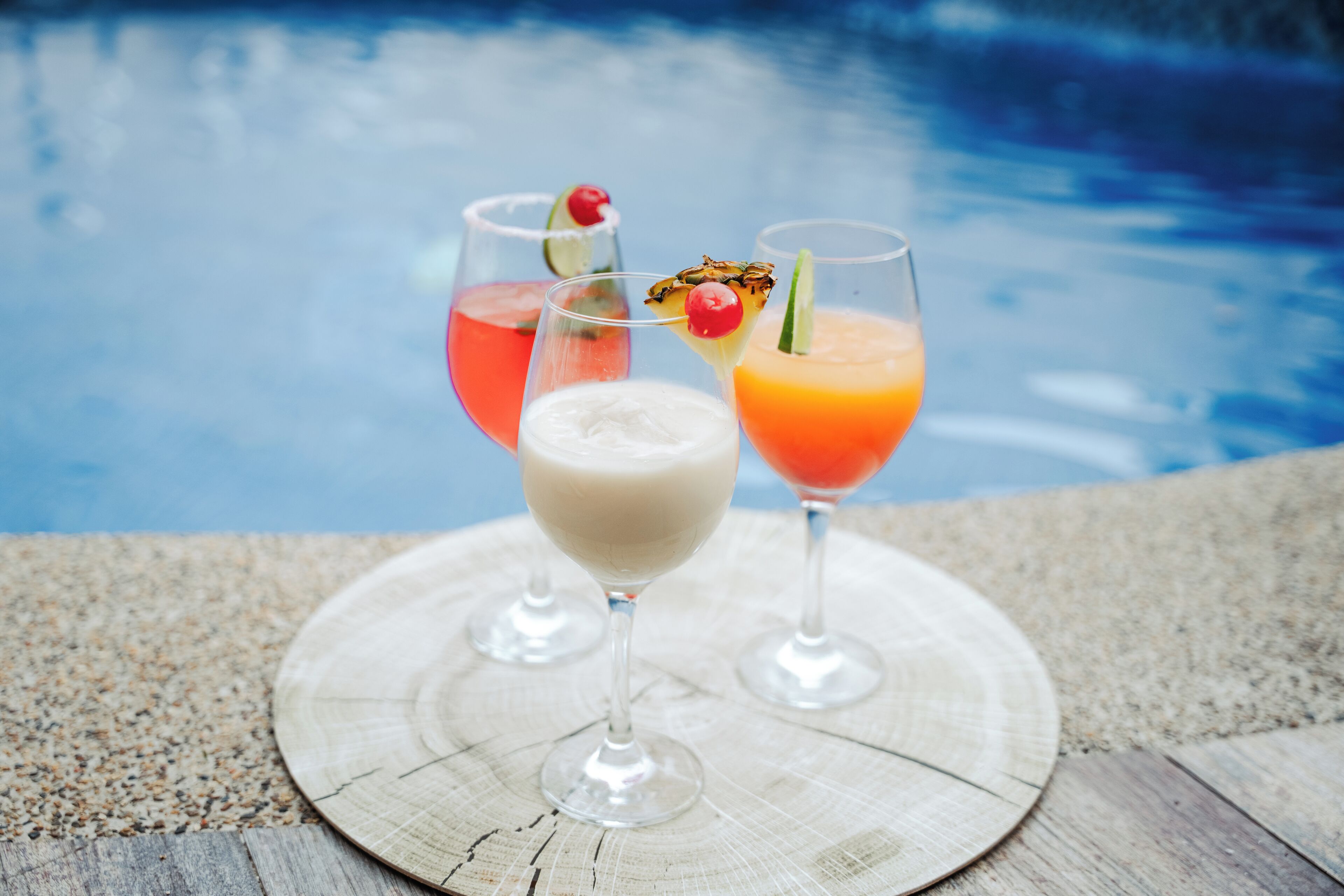Three cocktails on a wooden tray by a poolside.