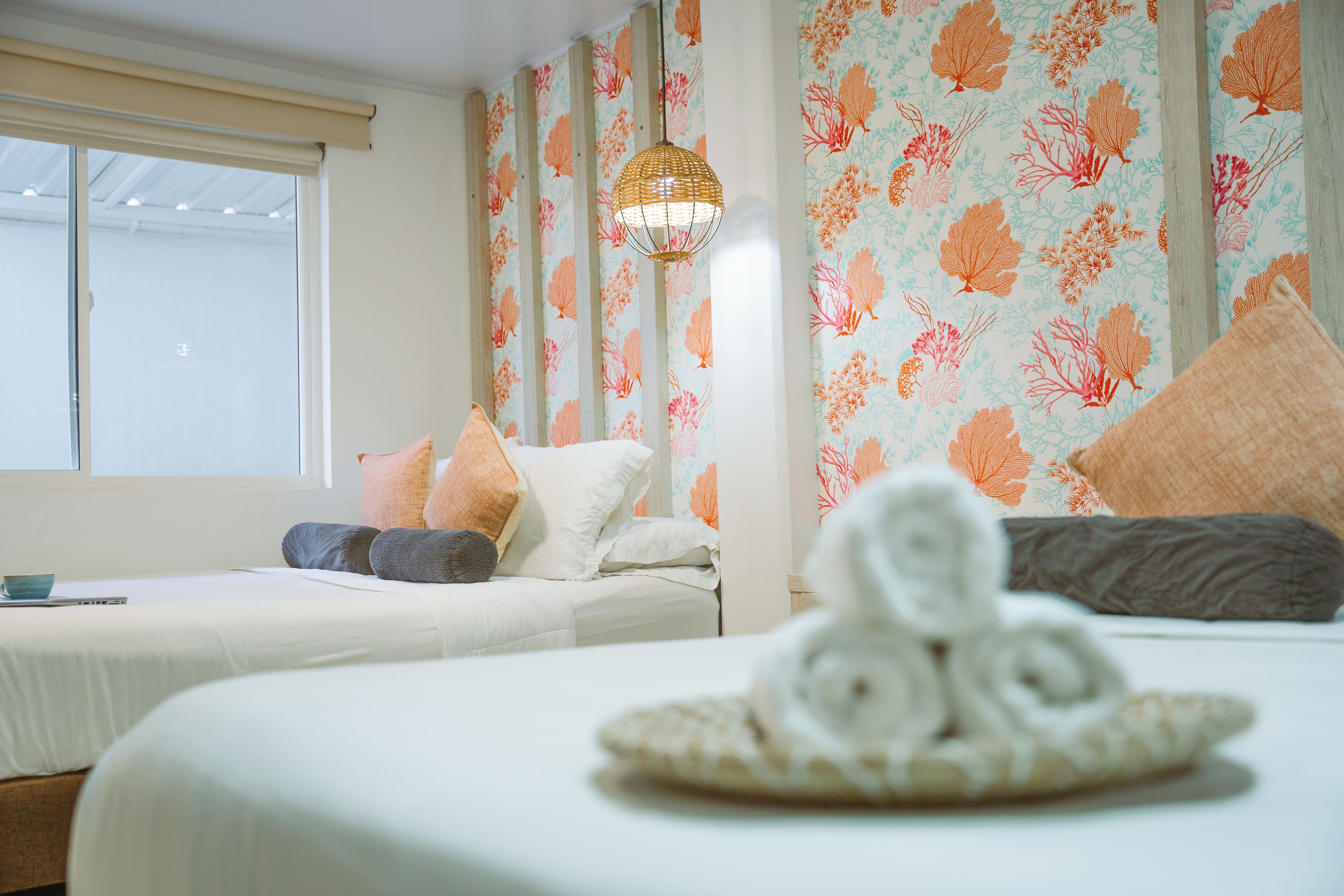 Une chambre d’hôtel avec deux lits, un papier peint à motifs floraux, un luminaire suspendu et des serviettes disposées sur le lit.