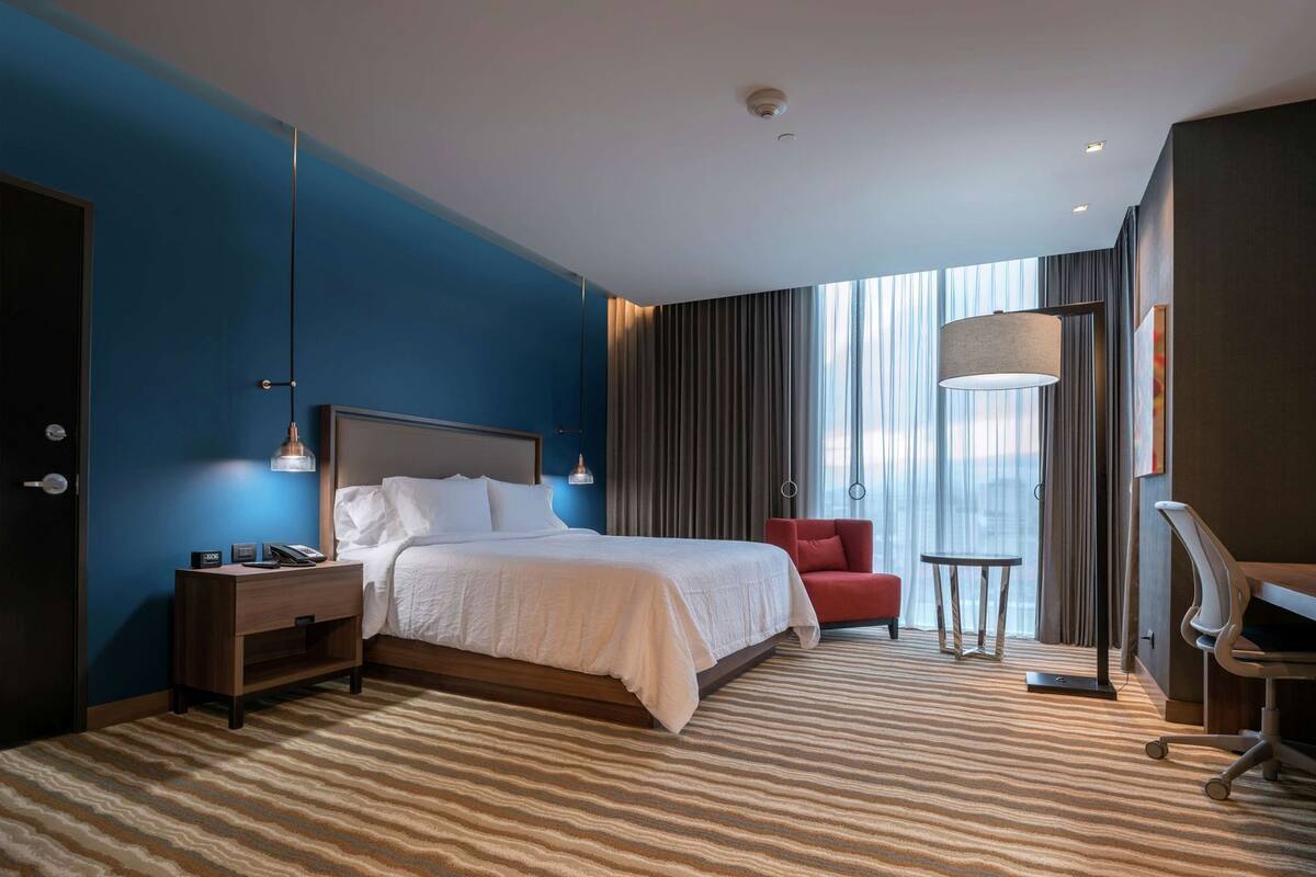 Habitación de hotel con cama, escritorio, silla, lámpara y vistas de la ciudad.