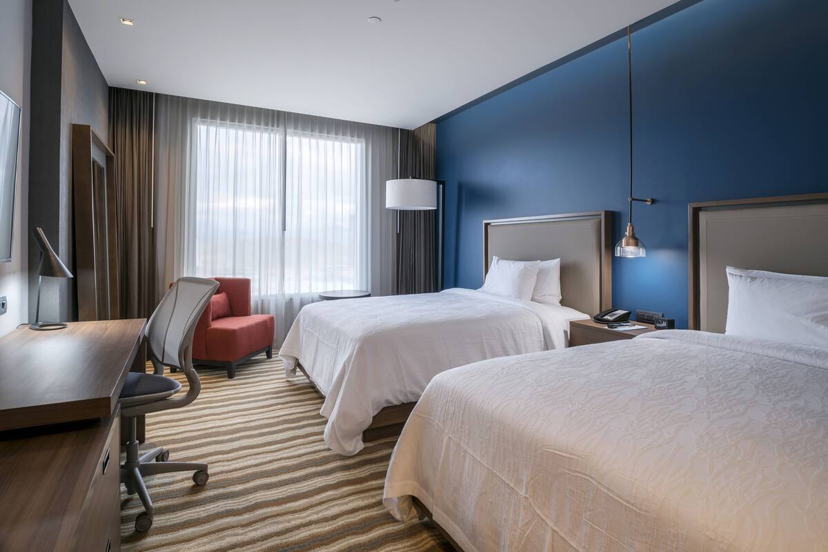 Habitación de hotel con dos camas, un escritorio, una silla, una lámpara y vistas al exterior.