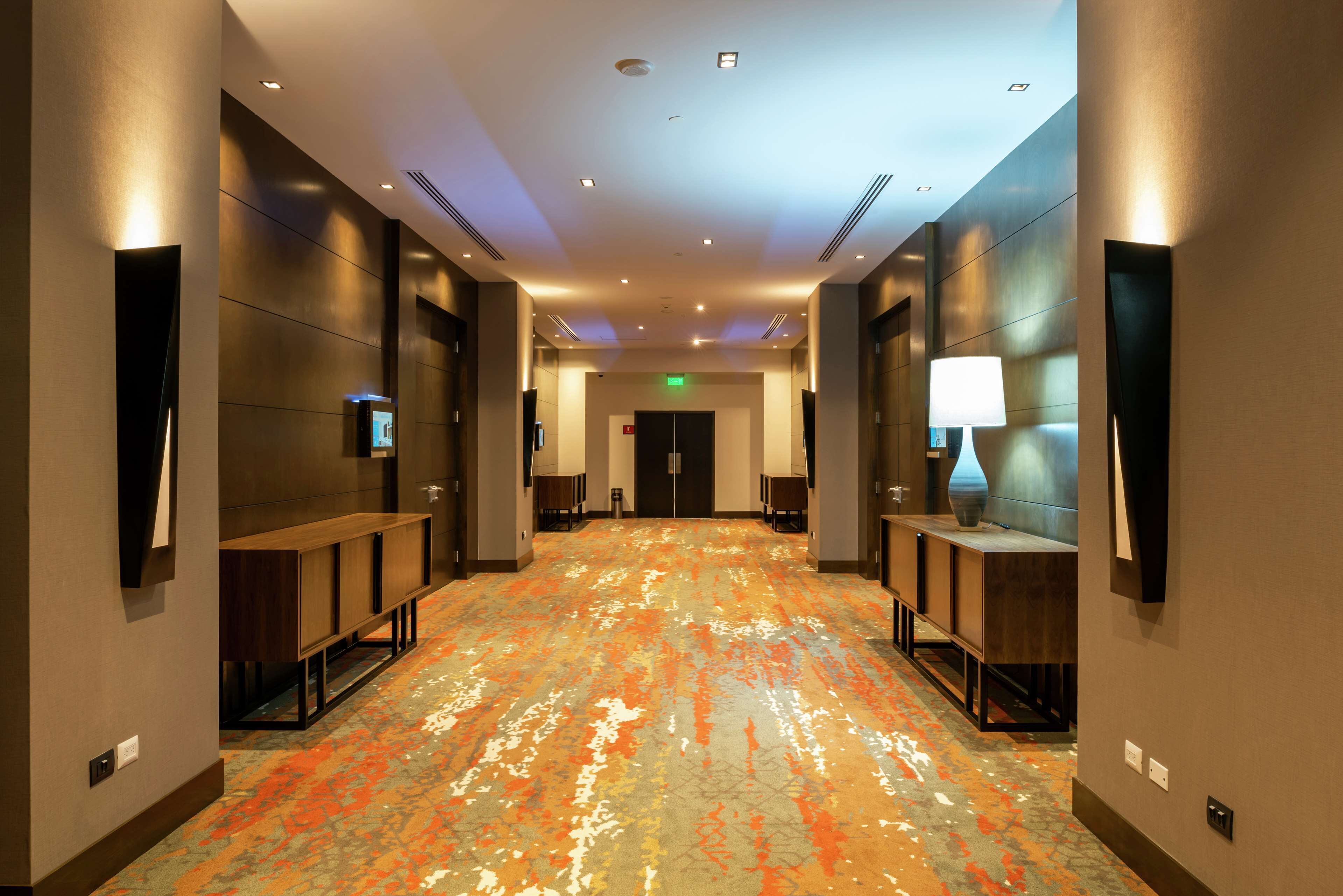 A hotel corridor with a patterned carpet, wooden benches, wall-mounted lamps, and a flat-screen TV.