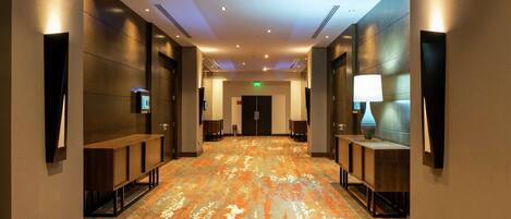 A hotel corridor with a patterned carpet, wooden benches, wall-mounted lamps, and a flat-screen TV.