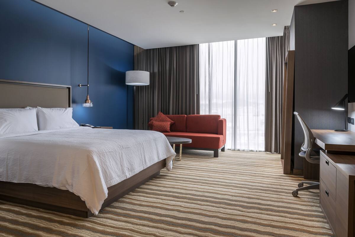 Una habitación de hotel con cama, escritorio, sofá rojo y un ventanal con cortinas.