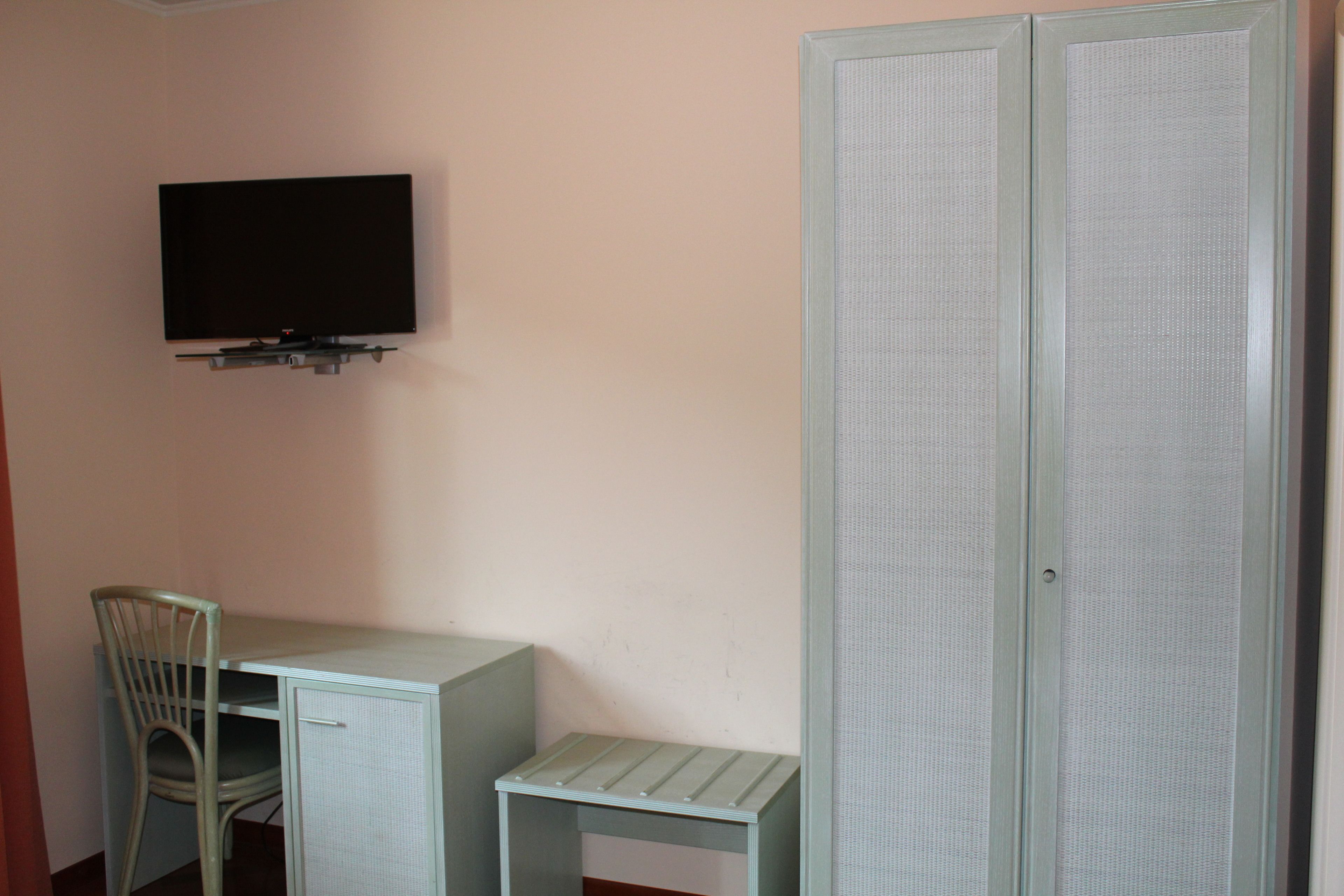Ein Zimmer mit Schreibtisch, Stuhl, Wandfernseher und Kleiderschrank.