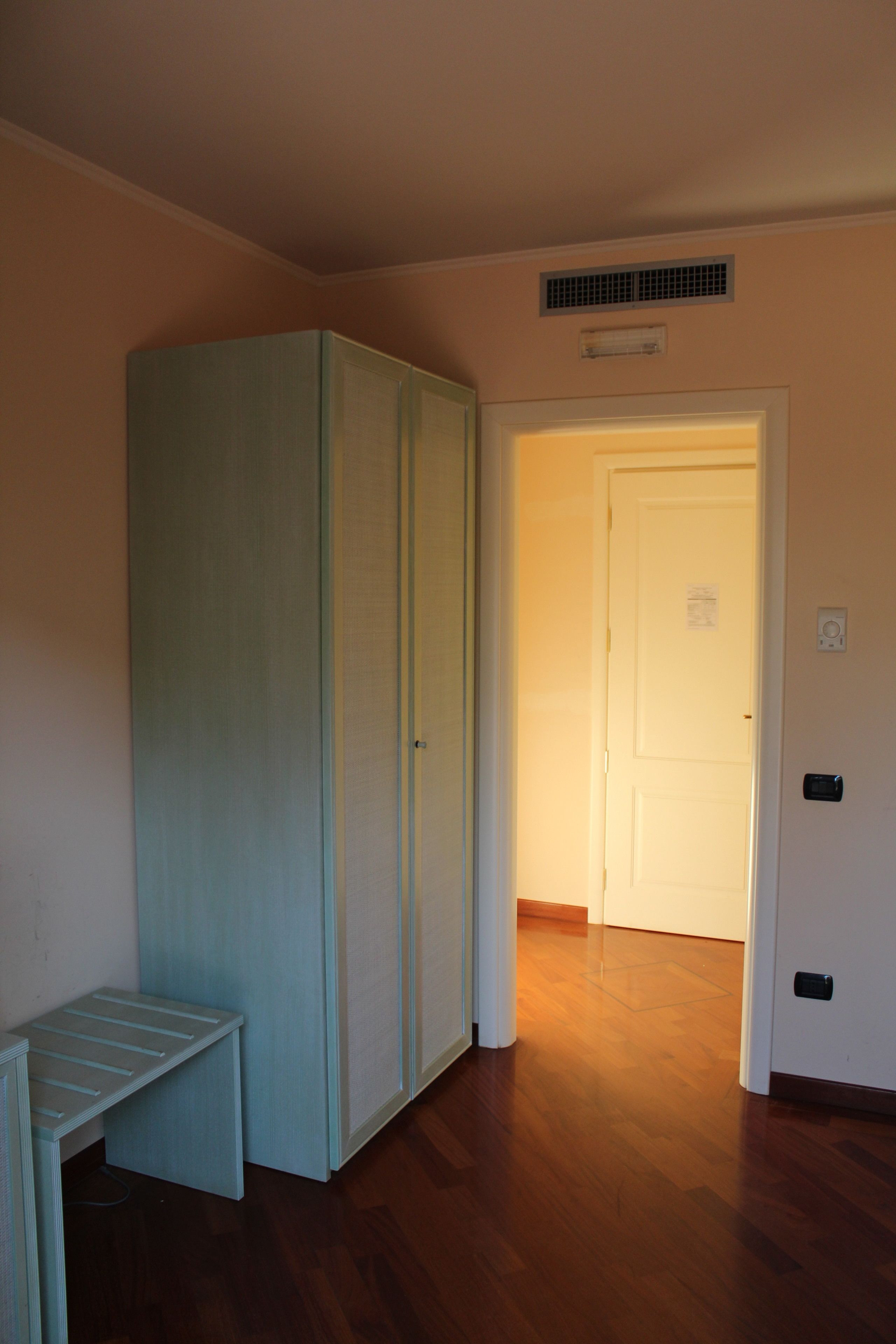Ein Zimmer mit Holzboden, einer Bank, einem Kleiderschrank und einer Tür zu einem weiteren Raum.