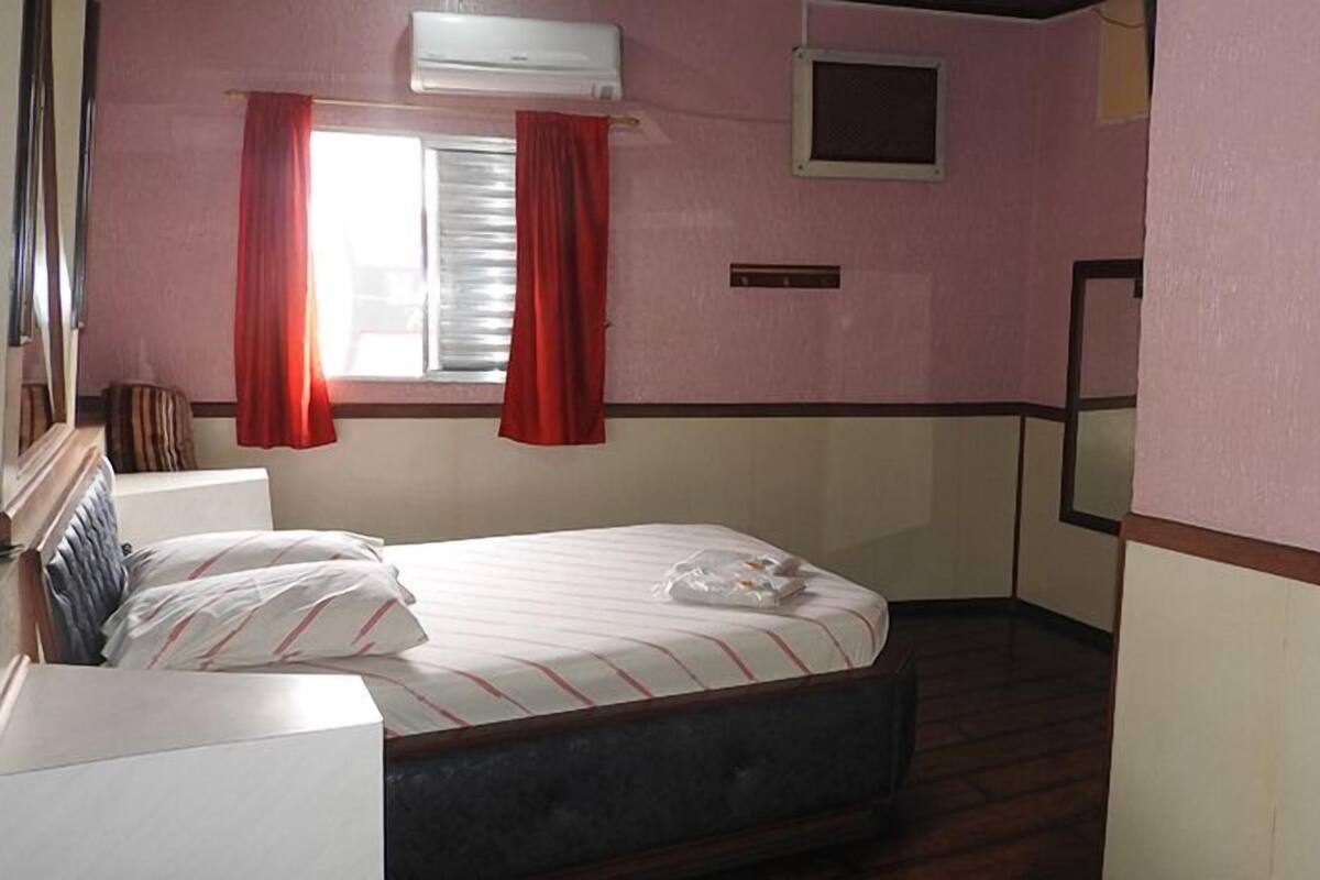 Une petite chambre avec un lit simple, une tête de lit en bois, une fenêtre avec des rideaux rouges, un climatiseur fixé au mur et une petite étagère.