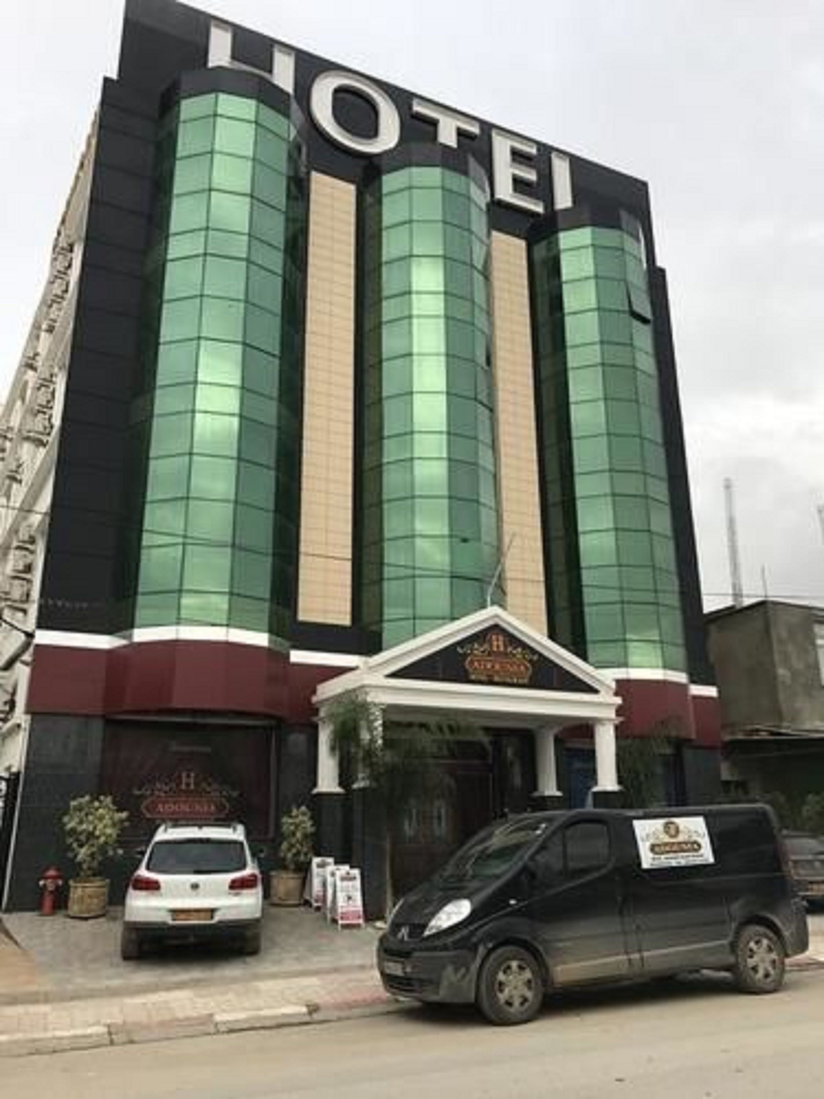 A hotel with a prominent 'HOTEL' sign on the facade, featuring three vertical glass windows and a central entrance with a decorative archway.