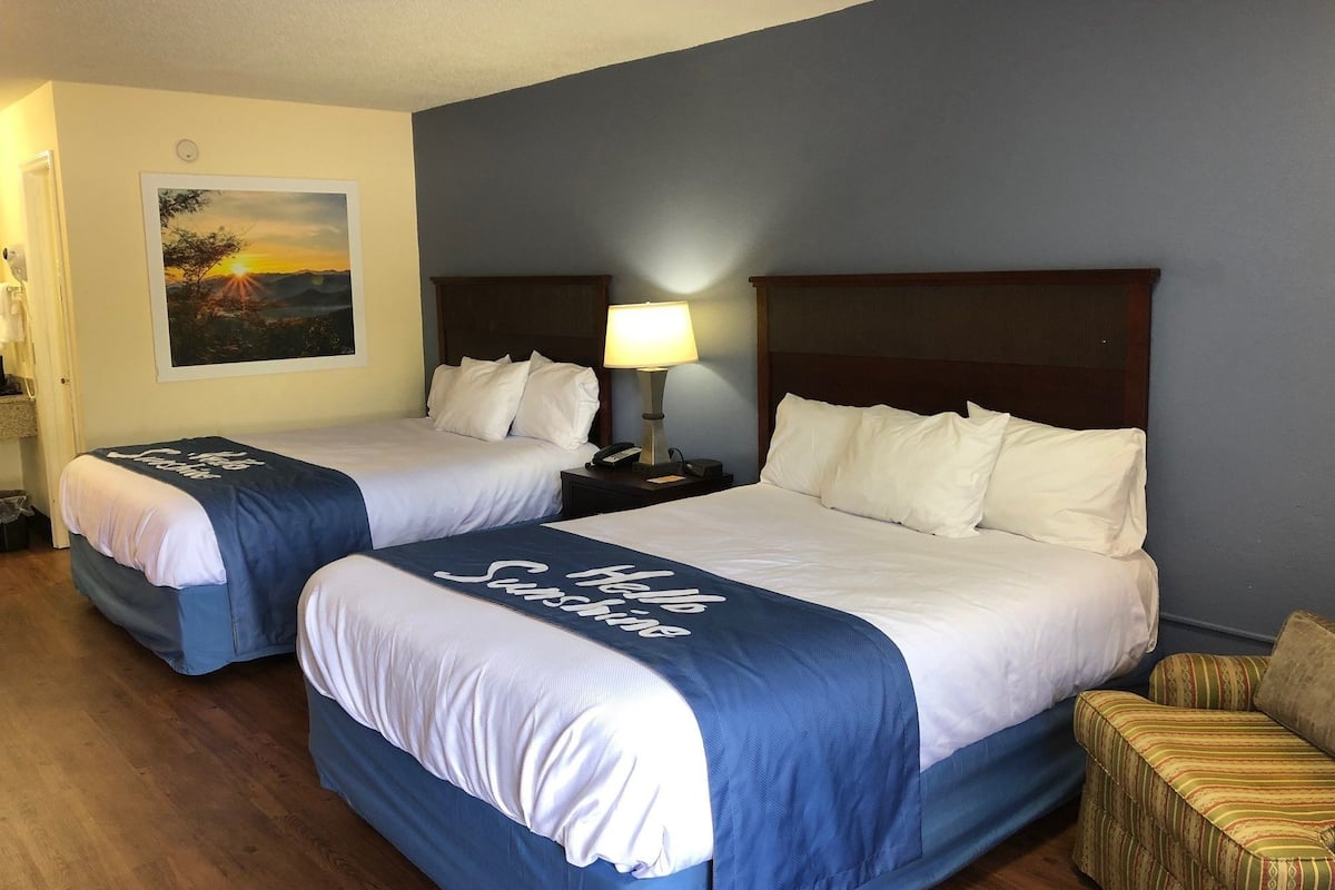 A hotel room with two beds, a nightstand with a lamp, a painting on the wall, and a small seating area.