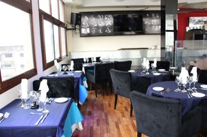 Un restaurante con mesas dispuestas para comer, una pantalla de televisión en la pared y vistas de la ciudad a través de grandes ventanales.