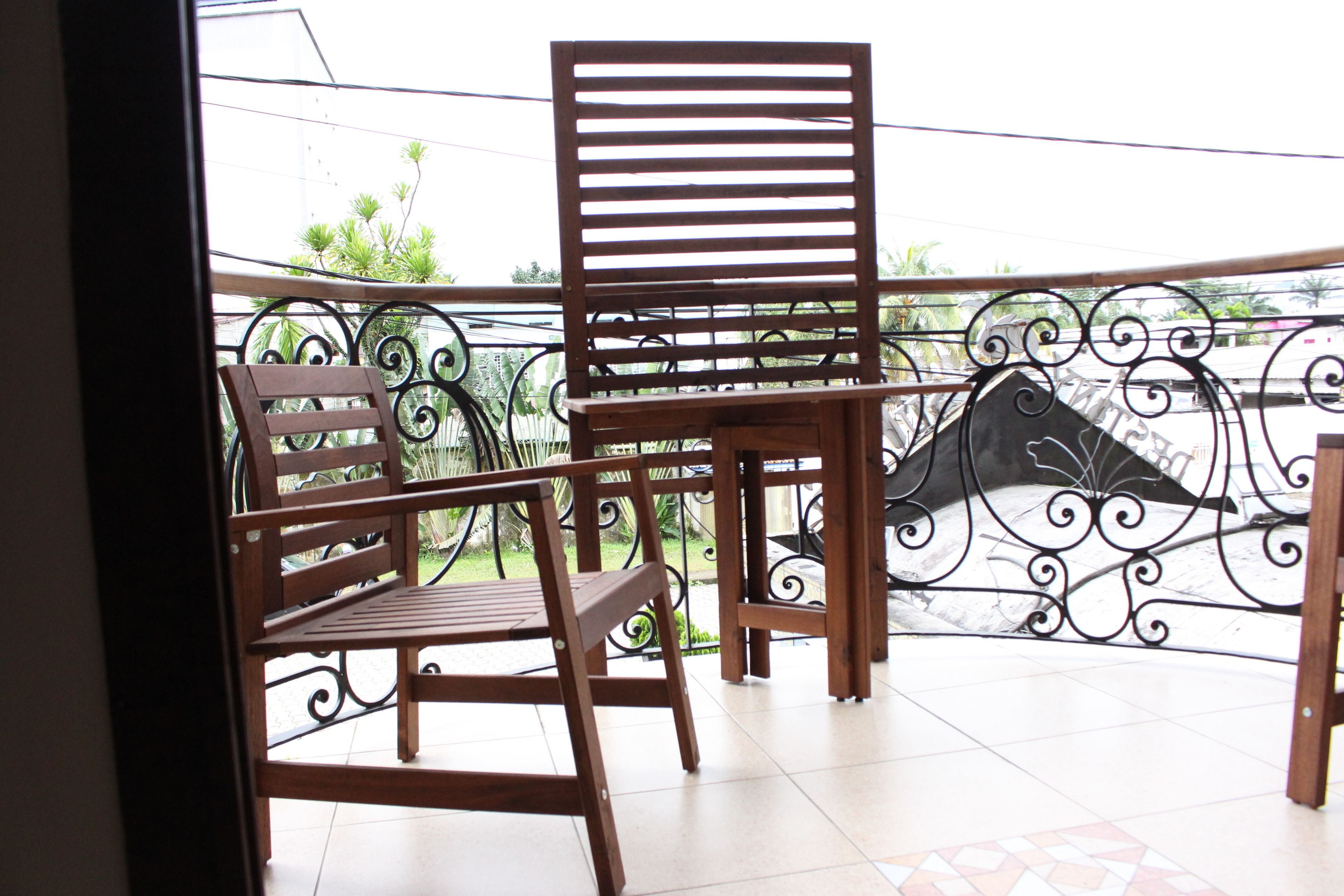 A balcony with two wooden chairs and a table, featuring a decorative metal railing and a view of greenery.