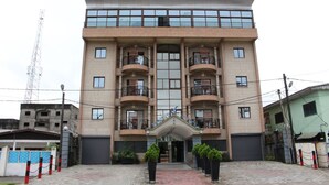 A multi-story hotel with a central entrance, balconies on each floor, and a parking area in front.