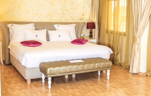 Una cama bien tendida con sábanas blancas y dos almohadas rosas, una mesita de noche con una lámpara, y un piso de madera.