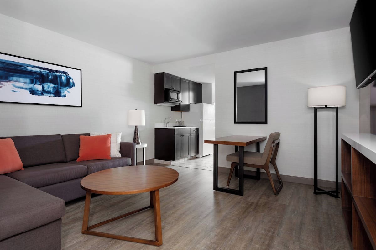 A modern living room with a sofa, coffee table, and a dining area with a table and chairs. There is a kitchen area with cabinets and a microwave.