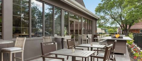 An outdoor patio with tables and chairs, large windows, and a fire feature.
