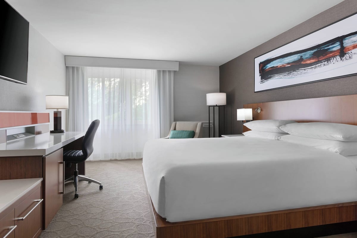 A hotel room with a large bed, a desk with a chair, a TV mounted on the wall, and a window with sheer curtains.