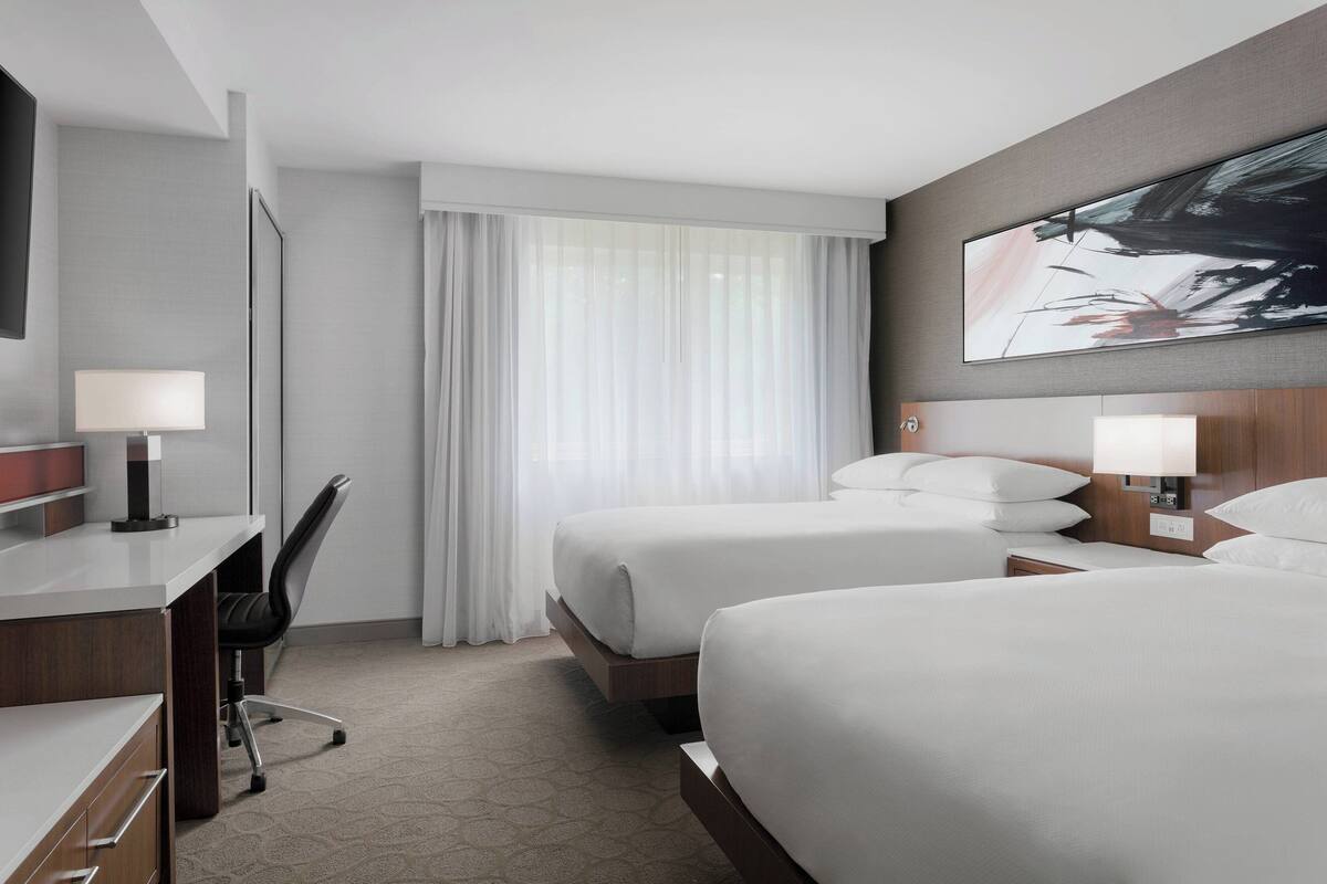 A hotel room with two beds, a desk, a chair, a large window with sheer curtains, and a painting on the wall.