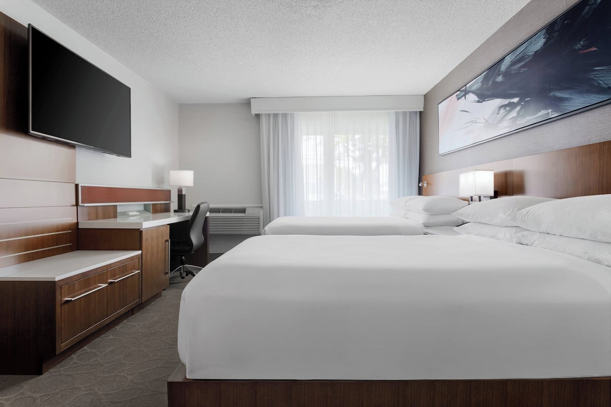 A hotel room with two beds, a desk, a TV, and a large window with curtains.