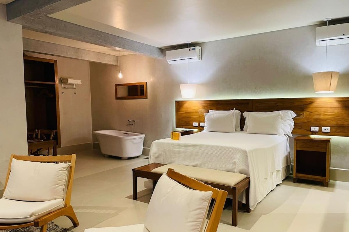 Une chambre d’hôtel comprenant un lit, deux tables de chevet, une commode, une chaise, une petite table, un canapé, une salle de bain avec une baignoire et une porte en bois.