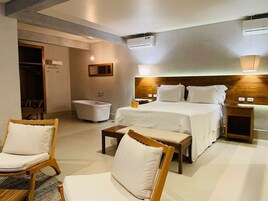 Een hotelkamer met een bed, twee nachtkastjes, een stoel, een kleine tafel, een bank, een badkamer met een badkuip en een houten deur.